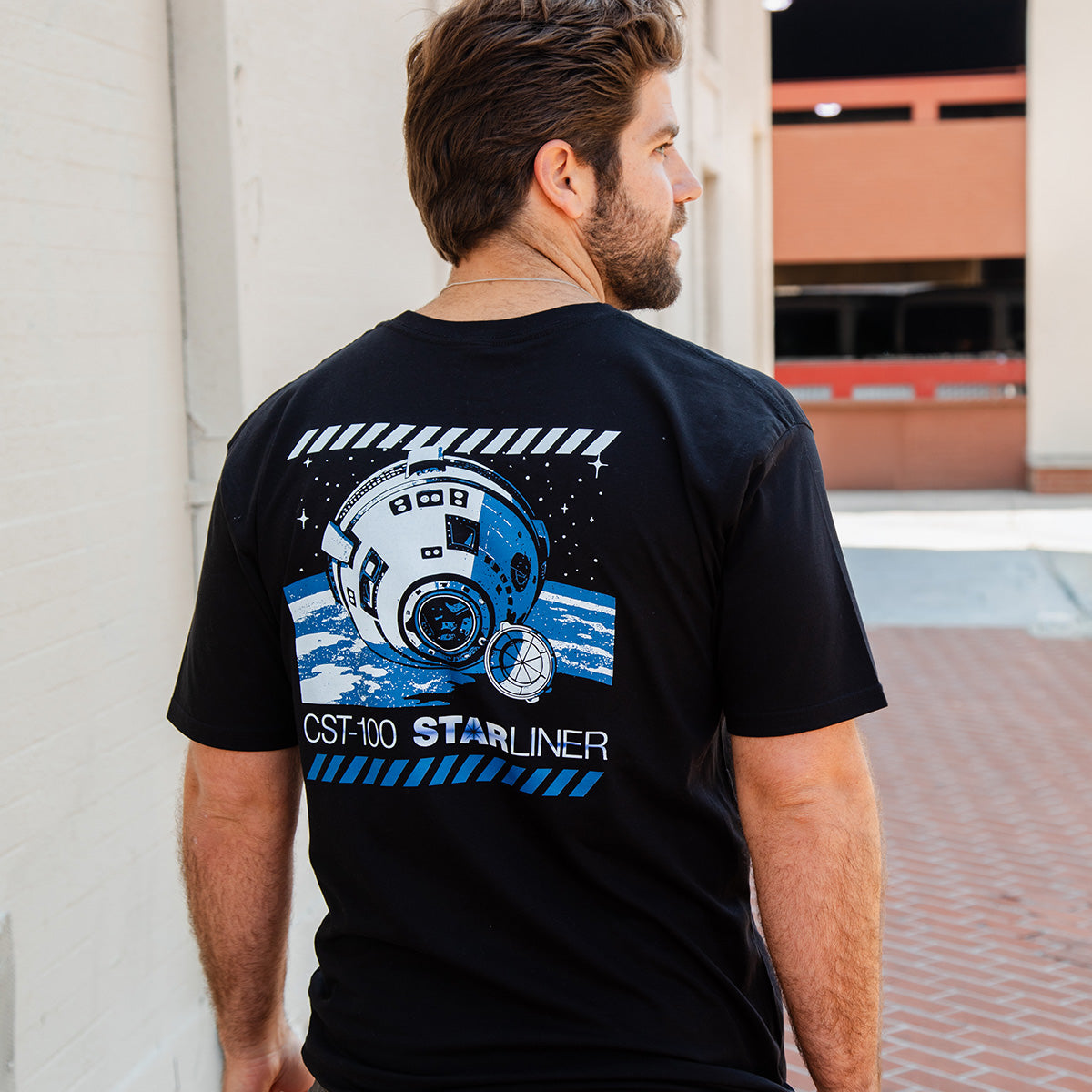 A man with brown hair and a beard wears the Boeing CST-100 Starliner Unisex Graphic T-Shirt by BOEING, featuring the spacecraft in space above Earth and CST-100 STARLINER text on the back to celebrate human spaceflight.