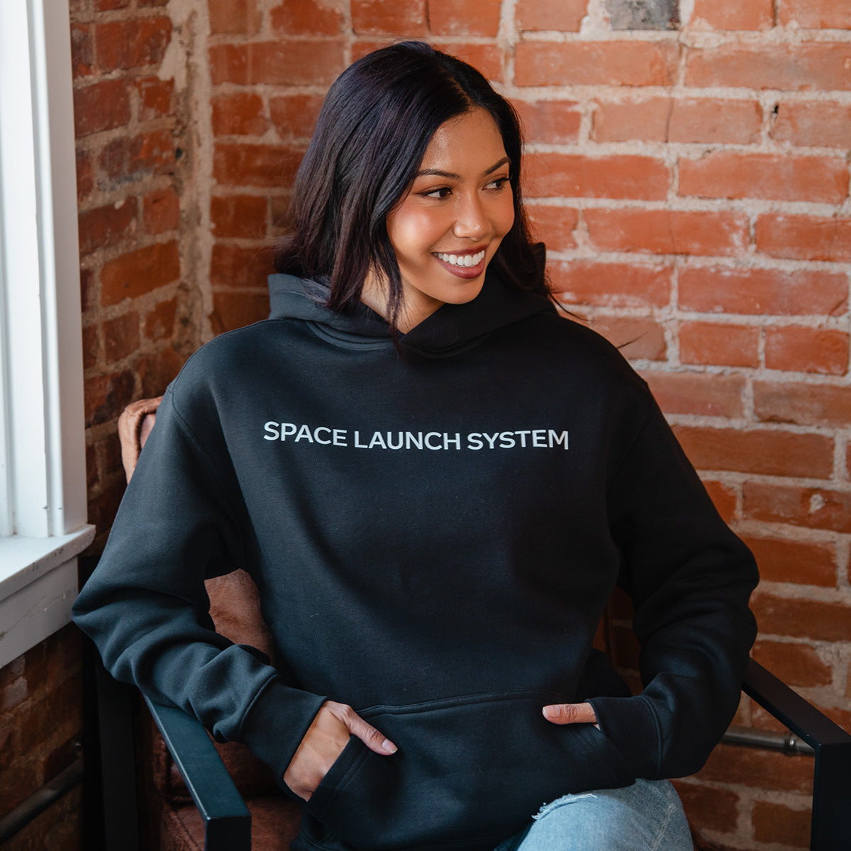 A woman with long dark hair smiles while sitting against a brick wall, wearing the BOEING SLS Artemis II Unisex Graphic Hoodie, featuring "SPACE LAUNCH SYSTEM" in white letters and celebrating the Artemis II mission.