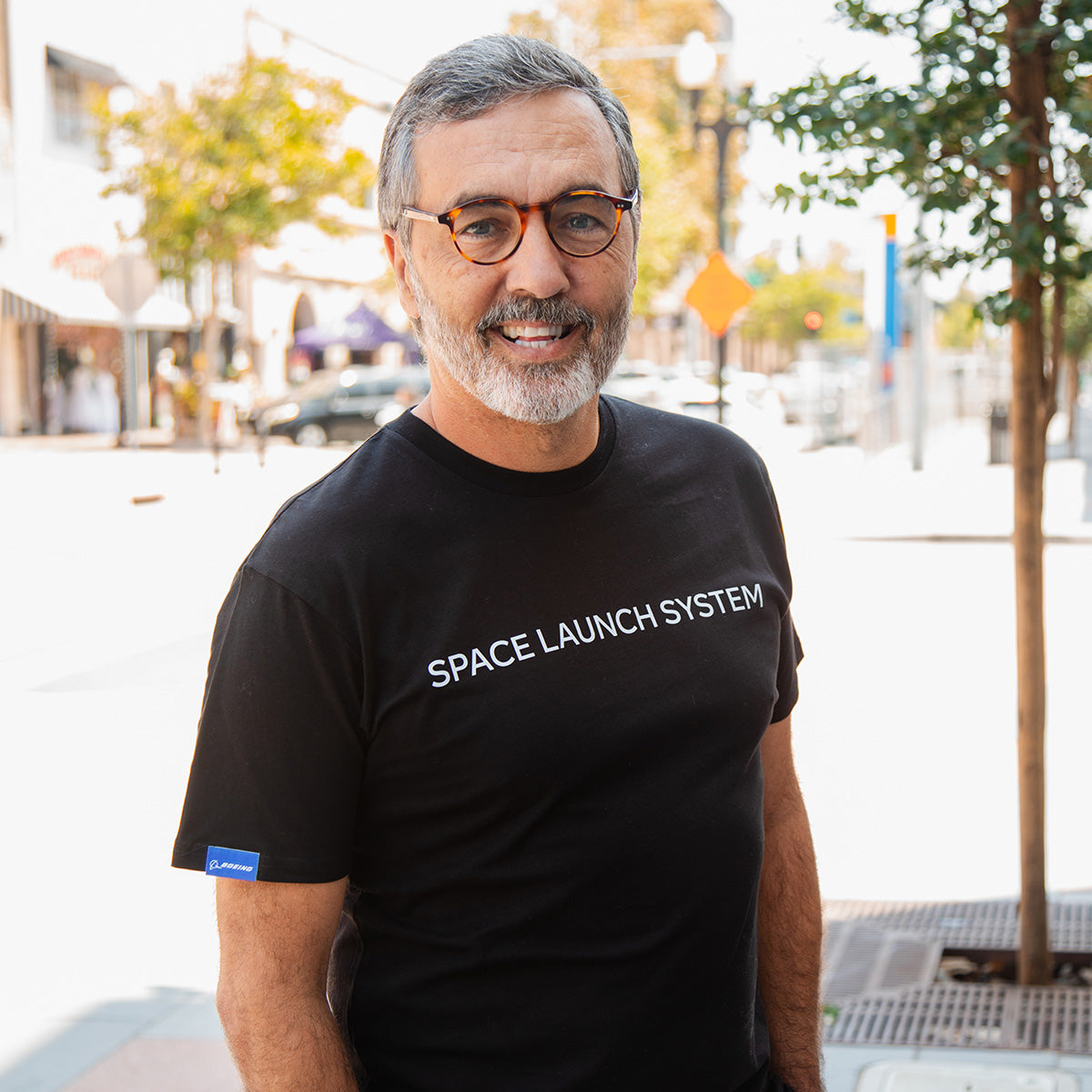 A man with gray hair and glasses smiles while standing outdoors in a black BOEING SLS Artemis II Unisex Graphic T-Shirt, near trees and buildings on a sunny city sidewalk—a proud moment for space fans.