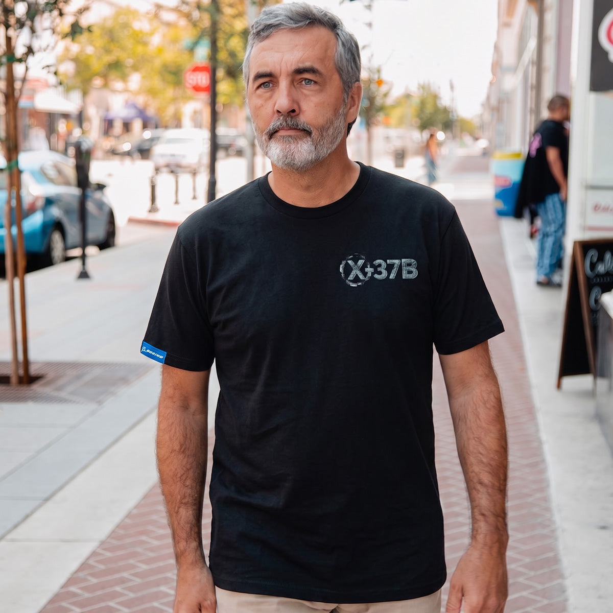 A middle-aged man with gray hair and a beard, likely a Boeing fan or space enthusiast, walks along a bright city sidewalk wearing a BOEING X-37B Orbital Test Vehicle Unisex Graphic T-Shirt.