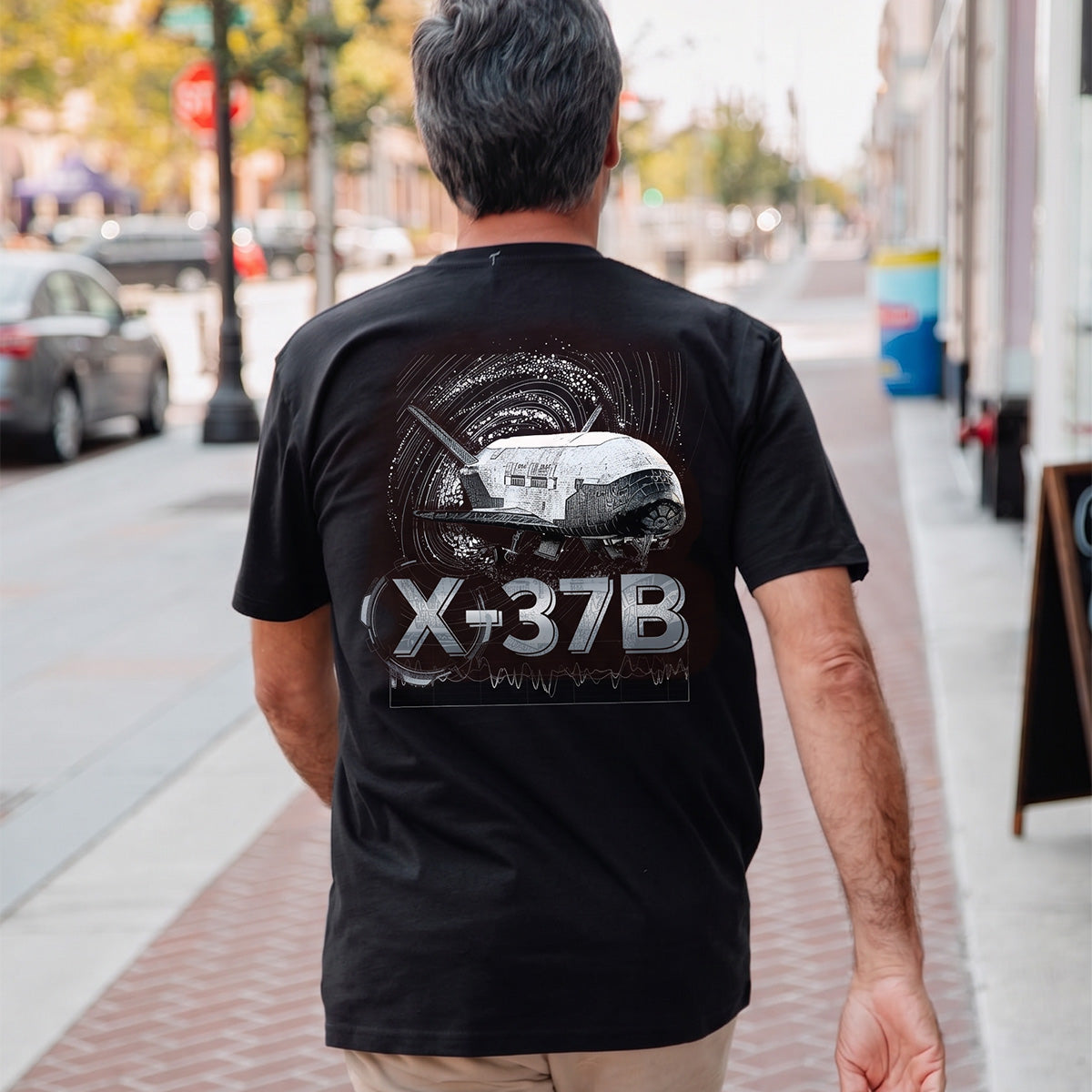 A man with gray hair walks down a city sidewalk in a Boeing X-37B Orbital Test Vehicle Unisex Graphic T-Shirt by BOEING, making it the perfect standout for space fans. Cars and trees line the street as he displays his love for aerospace.