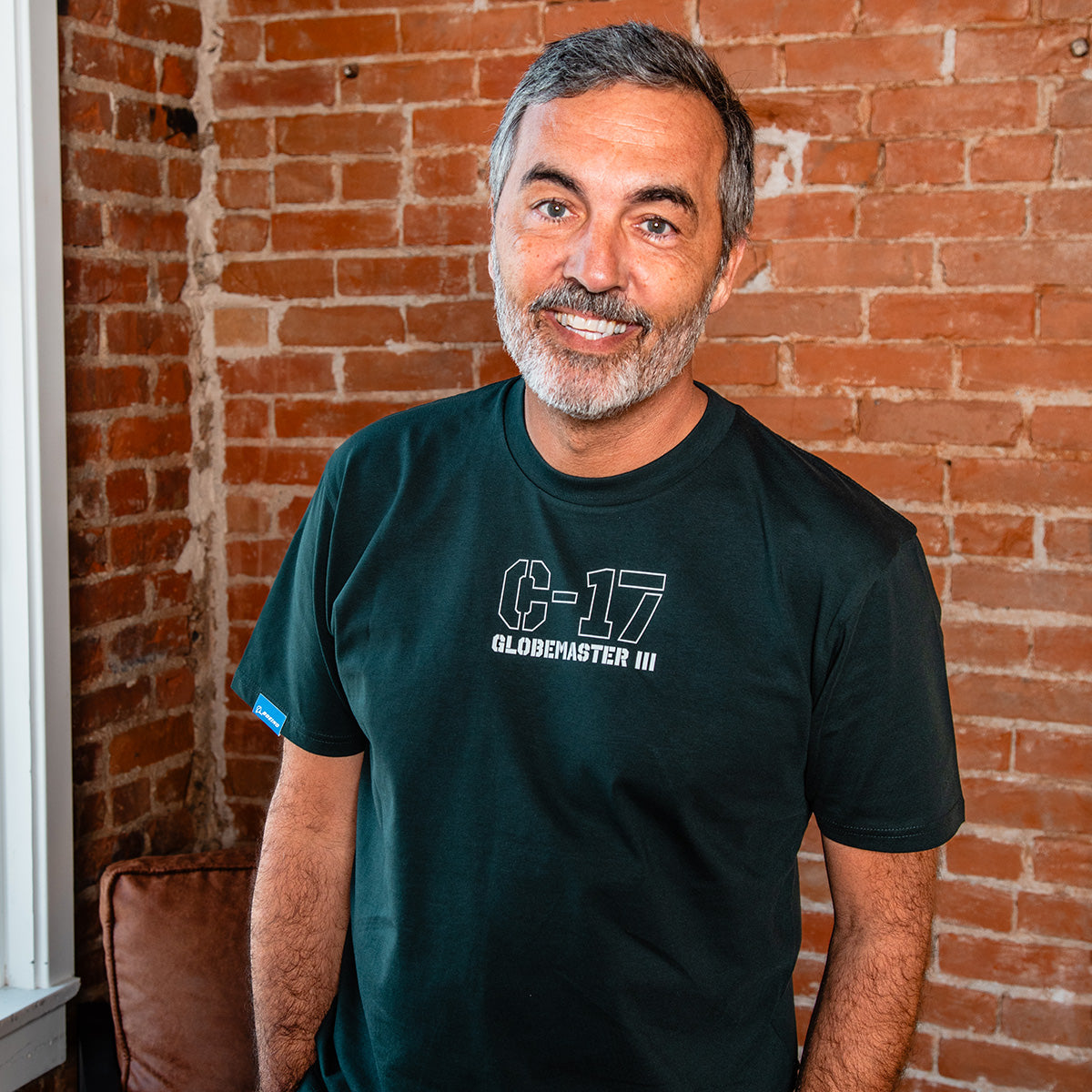 A smiling man with short gray hair and a beard stands indoors by a brick wall, wearing the BOEING C-17 Globemaster III Unisex Graphic T-Shirt.
