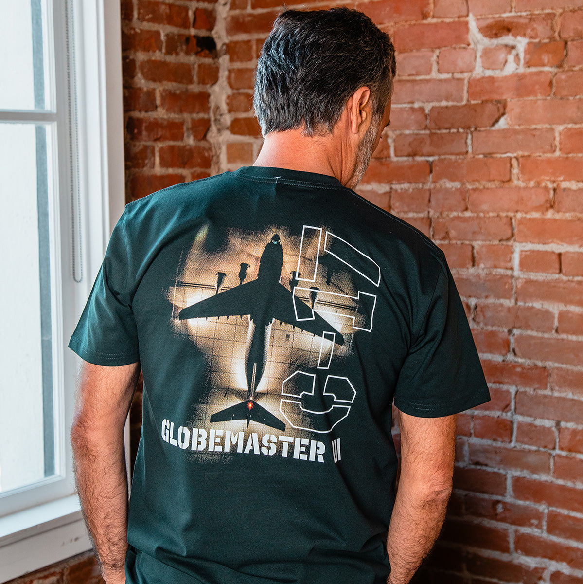 A man stands indoors facing a brick wall, wearing the BOEING C-17 Globemaster III Unisex Graphic T-Shirt featuring a bold graphic and stylized lettering on the back.