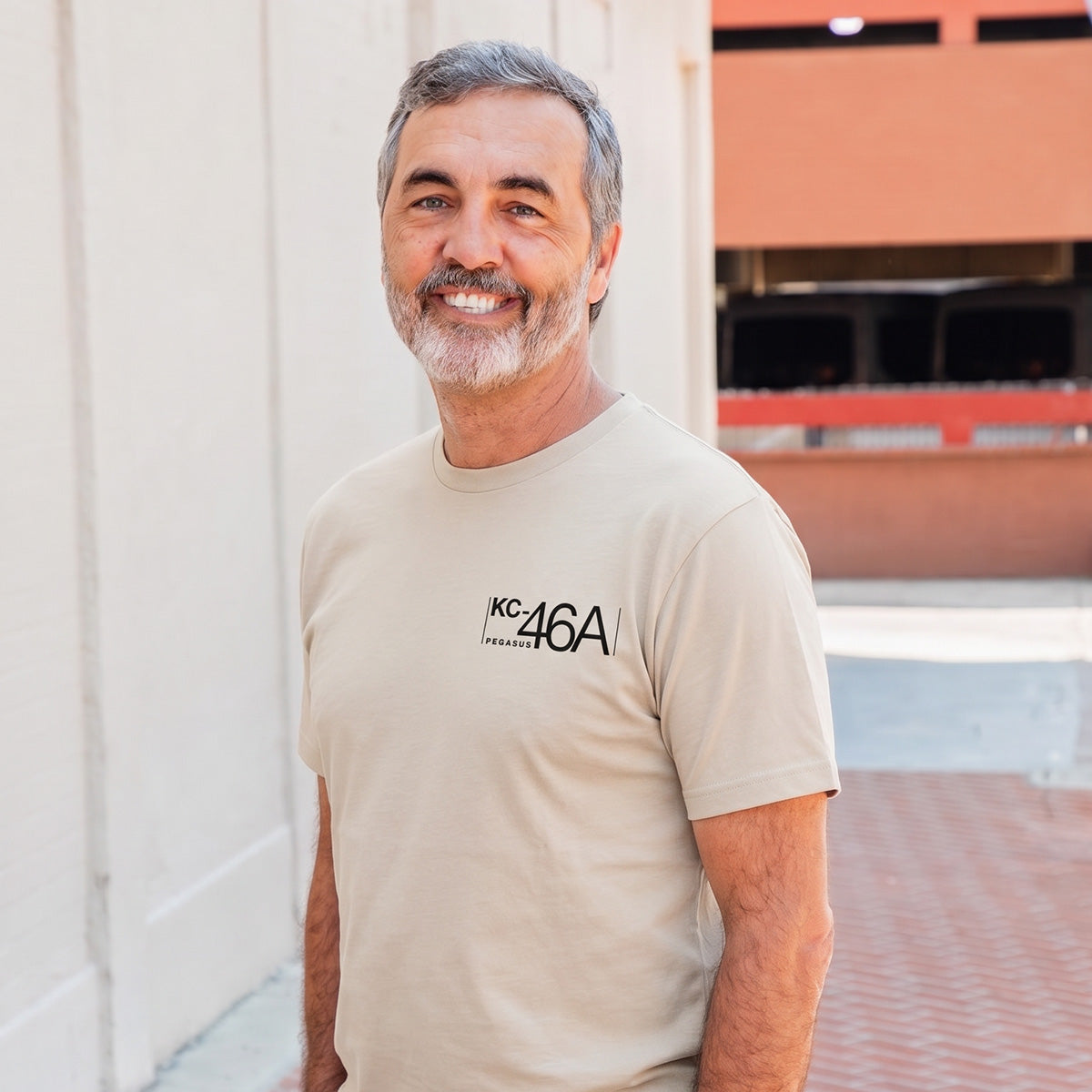 A middle-aged man with gray hair and a beard smiles outdoors in front of a light wall, wearing the BOEING KC-46A Pegasus Unisex Graphic T-Shirt—an ideal choice for aviation fans and Boeing supporters.