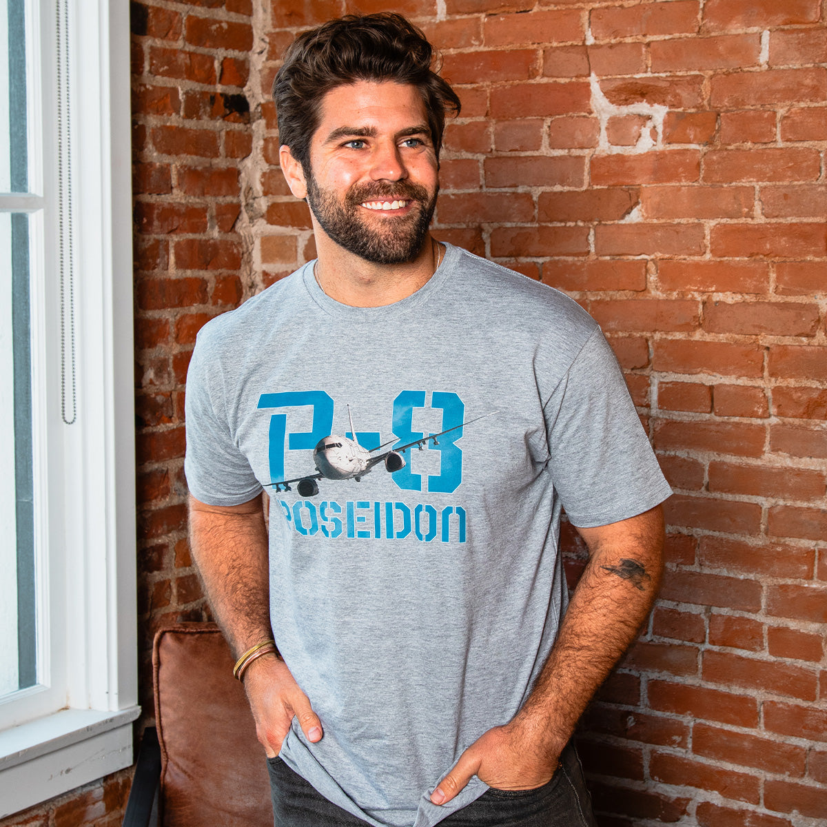 A bearded man with dark hair smiles indoors by a brick wall, wearing the BOEING P-8A Poseidon Unisex Graphic T-Shirt—gray with an airplane graphic and “P-8 Poseidon” text—ideal for aviation or Boeing fans.