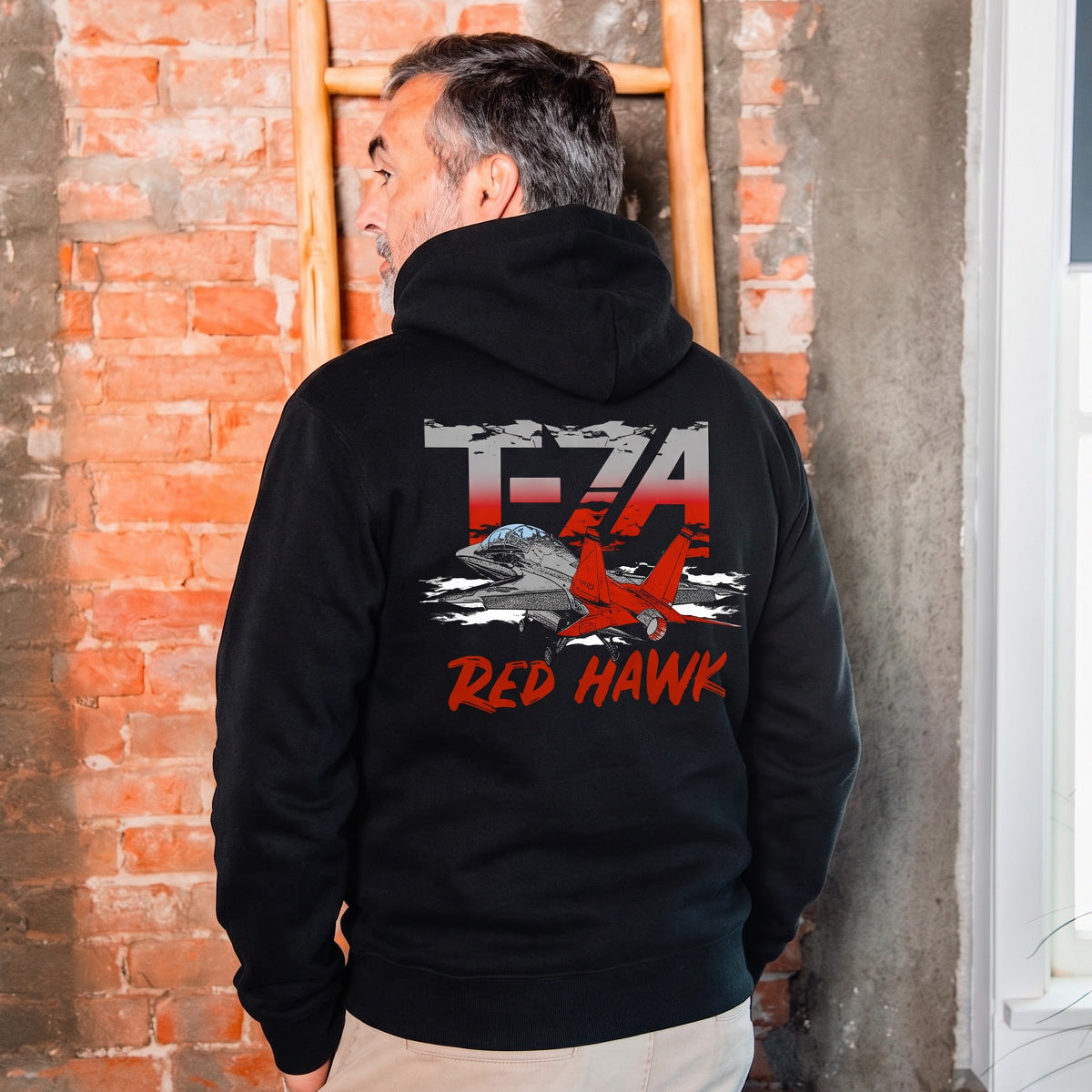 A man stands by a brick wall and wooden ladder wearing the BOEING T-7A Red Hawk Unisex Graphic Hoodie, featuring the T-7A Red Hawk jet, a soaring hawk, cloud graphics, and bold red and white T-7A Red Hawk text on the back.