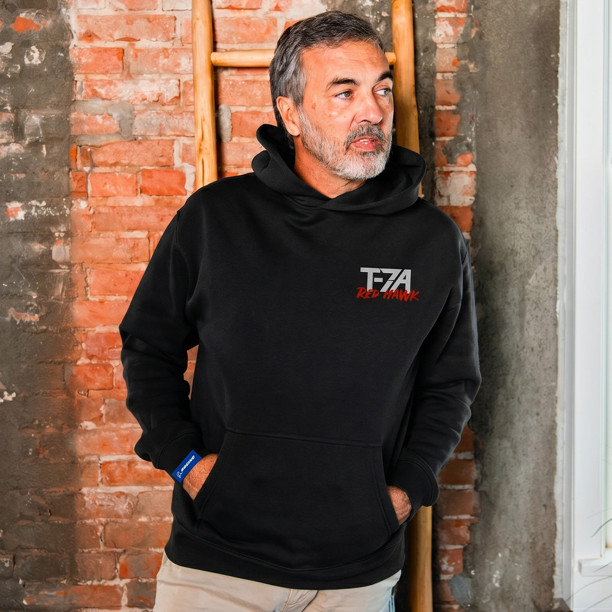 A man with gray hair and a beard wears a BOEING T-7A Red Hawk Unisex Graphic Hoodie, standing casually by a weathered brick wall, wooden ladder, and nearby window.