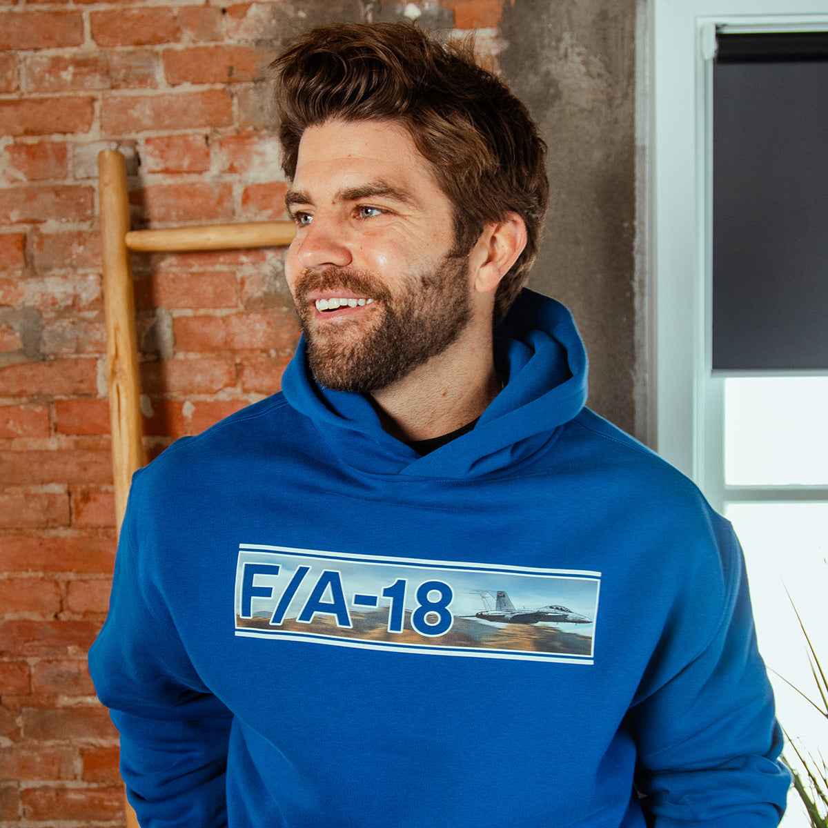 A smiling man with brown hair and a beard wears the BOEING F/A-18 Super Hornet Unisex Graphic Hoodie, perfect for aviation fans, standing indoors by a brick wall and window.