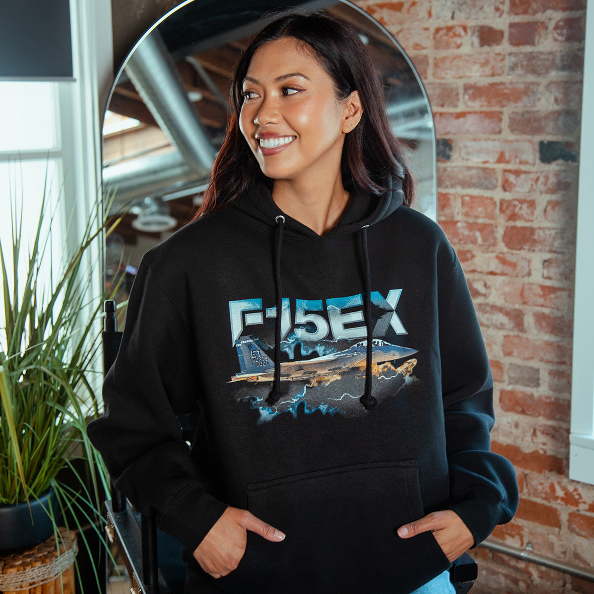 A smiling woman indoors by a brick wall and mirror wears the BOEING F-15EX Eagle II Unisex Graphic Hoodie, featuring a fighter jet graphic—ideal for aviation fans and Boeing supporters. Green plants are visible in the background.