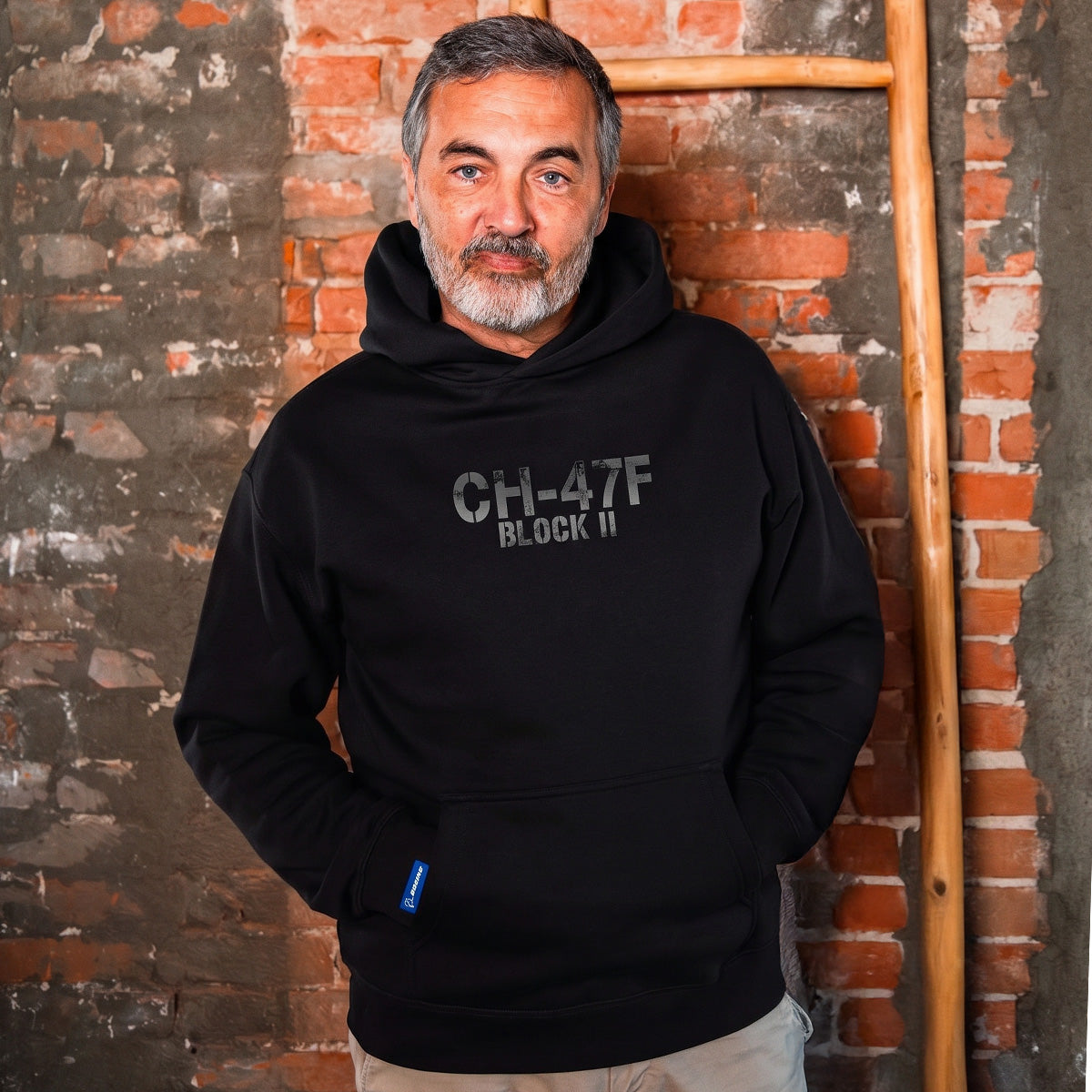 A man with gray hair and a beard wears a black BOEING CH-47F Block II Unisex Graphic Hoodie, standing before a weathered brick wall and wooden ladder—an ideal choice for aviation fans and dedicated Boeing supporters.