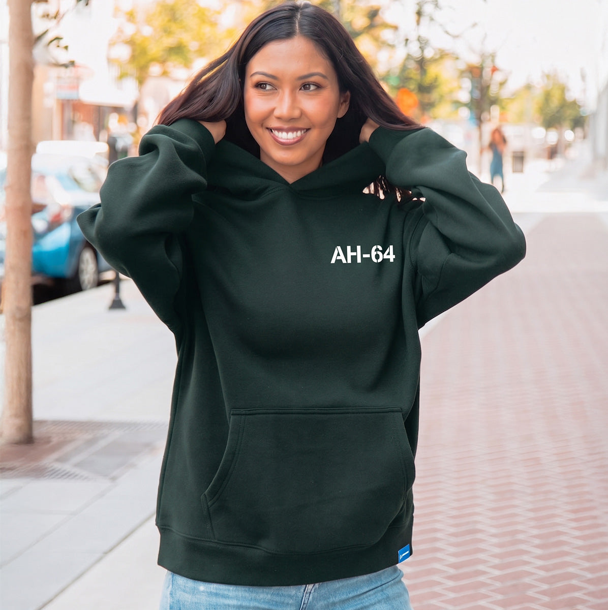 A woman smiles on a city sidewalk, wearing a dark green Boeing AH-64 Apache Unisex Graphic Hoodie by BOEING and holding the hood with both hands. Trees and parked cars line the street, creating a relaxed urban vibe.