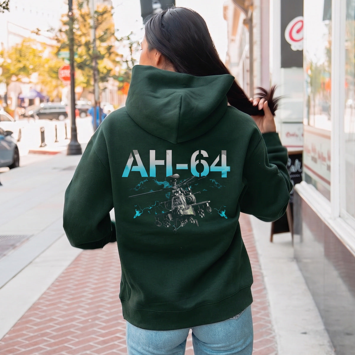 A person wearing the BOEING AH-64 Apache Unisex Graphic Hoodie, featuring an AH-64 graphic on the back, walks down a city sidewalk while holding their long hair with one hand.