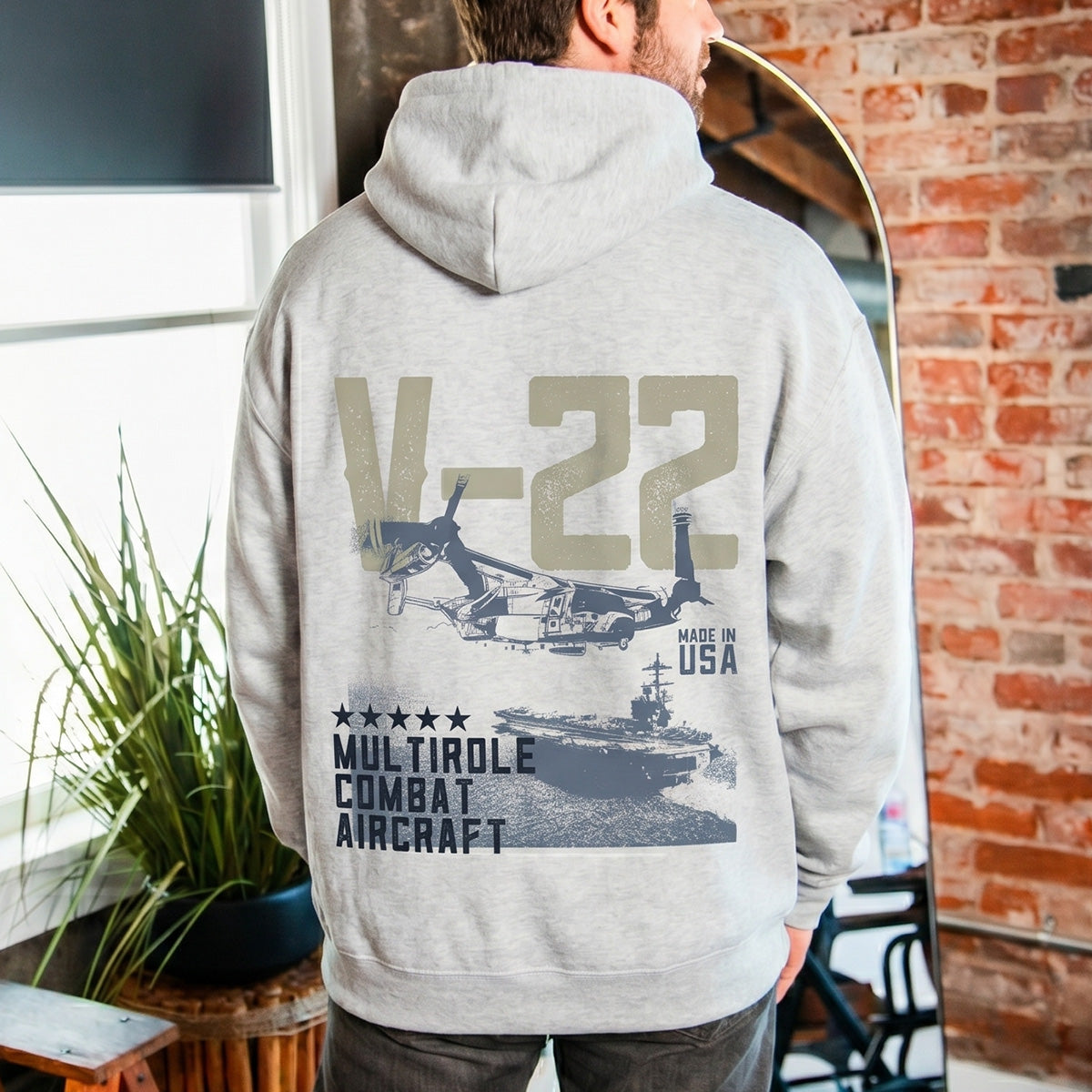 A man wears the BOEING V-22 Osprey Unisex Graphic Hoodie in gray, featuring a bold back graphic of the V-22 aircraft, stars, and “V-22 Multirole Combat Aircraft” plus “Made in USA”—celebrating Boeing innovation.