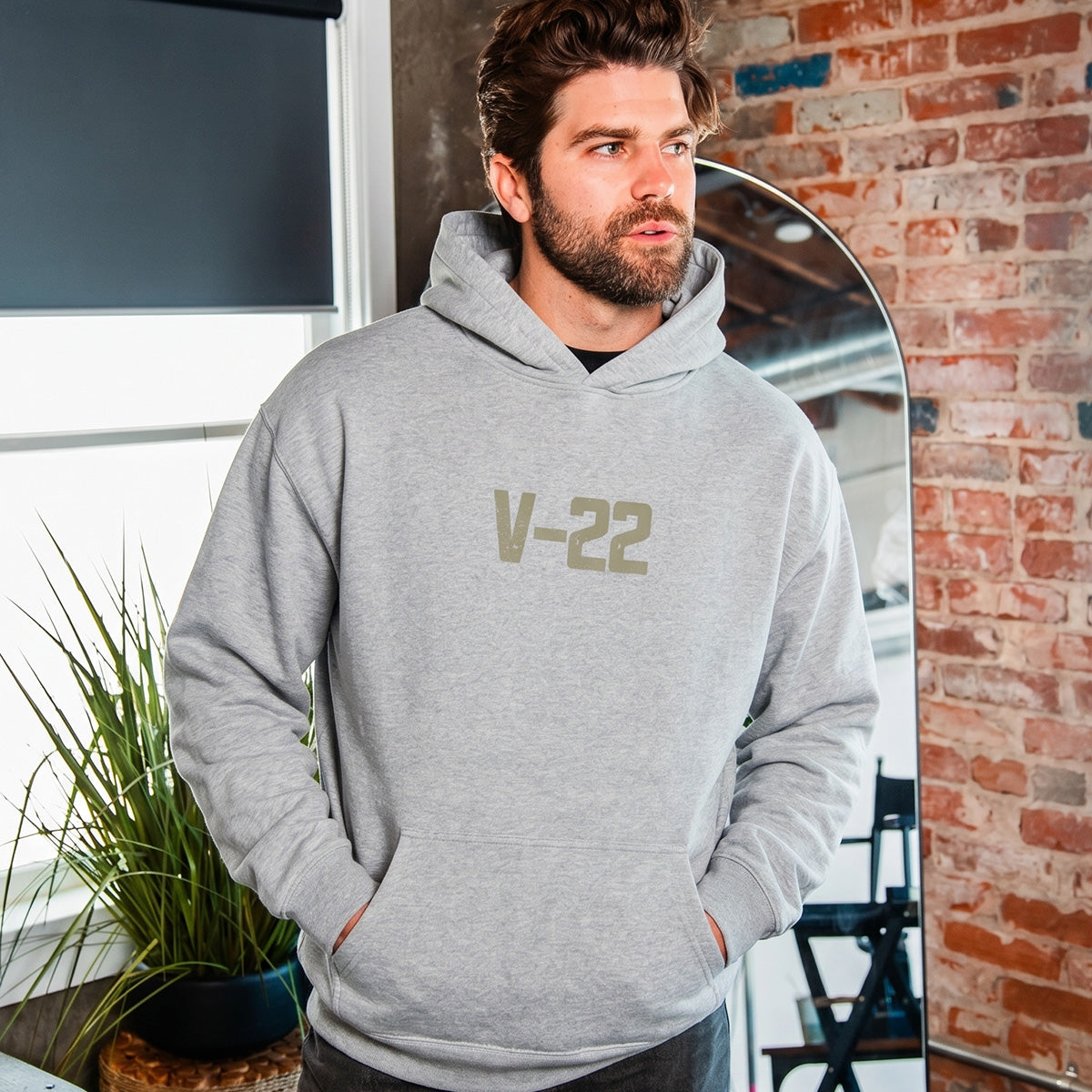 A man with brown hair and a beard wears the BOEING V-22 Osprey Unisex Graphic Hoodie indoors by a window, green plant, and brick wall, with his hands in the hoodie pockets.