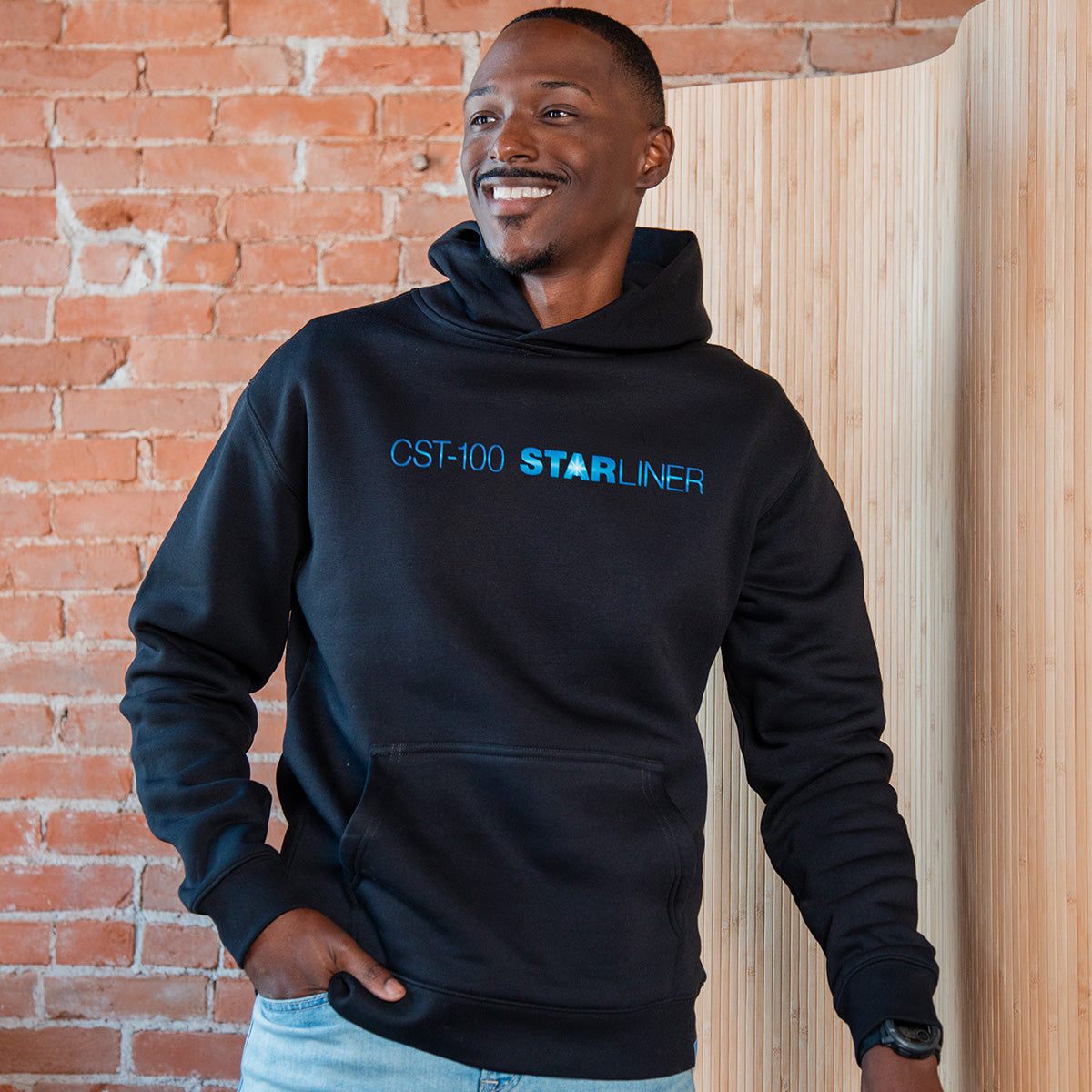 A man smiles in front of a brick wall and wood panel while wearing the BOEING CST-100 Starliner Unisex Graphic Hoodie, featuring blue text that celebrates human spaceflight achievements.
