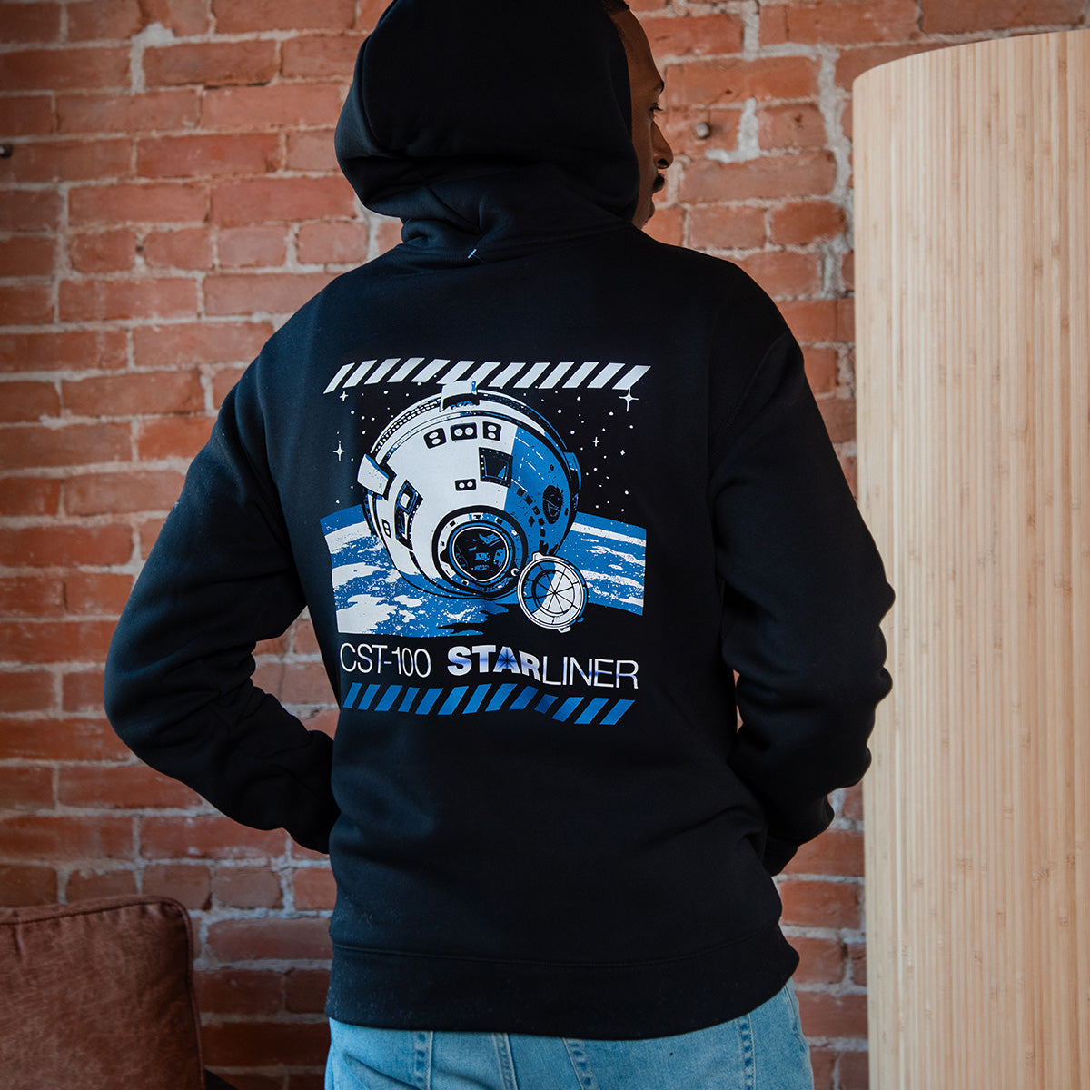 Wearing a BOEING CST-100 Starliner Unisex Graphic Hoodie with a space-themed design, someone stands facing away from the camera in front of a brick wall, displaying their passion for human spaceflight.