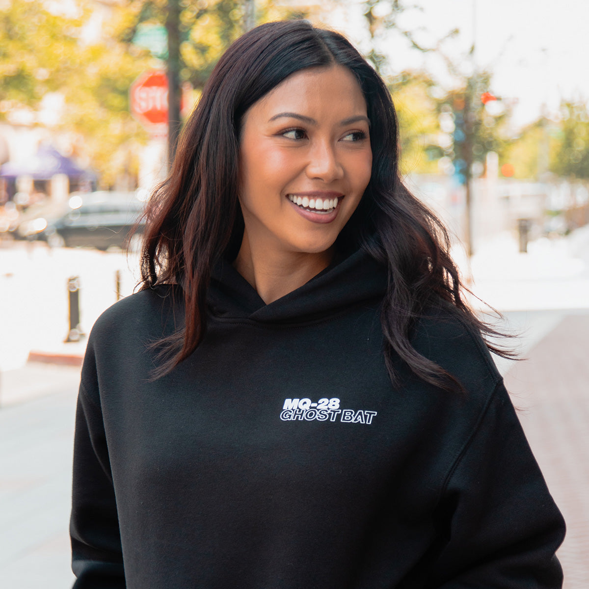 A woman with long dark hair smiles outdoors, wearing a BOEING MQ-28 Ghost Bat Unisex Graphic Hoodie. The black sweatshirt displays ‘MQ-28 Ghost Bat,’ referencing advanced unmanned flight. Trees and a blurred street are in the background.