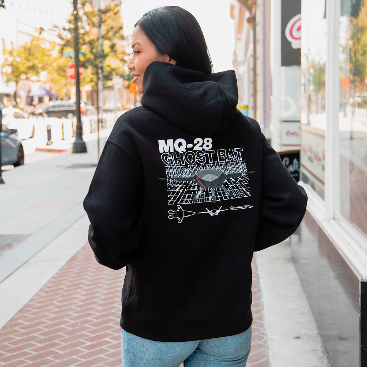 A person stands on a city sidewalk wearing a BOEING MQ-28 Ghost Bat Unisex Graphic Hoodie, featuring MQ-28 Ghost Bat graphics above a digital grid and aircraft designs, highlighting unmanned flight. The wearer faces away with hands in pockets.