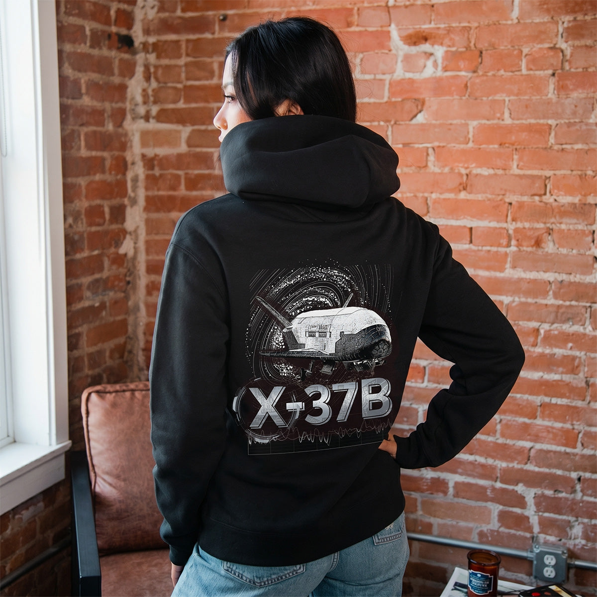Someone wears a Boeing X-37B Orbital Test Vehicle Unisex Graphic Hoodie by BOEING, featuring a white space shuttle design, while standing in a cozy room with exposed brick and a brown chair—a stylish tribute to spaceflight.