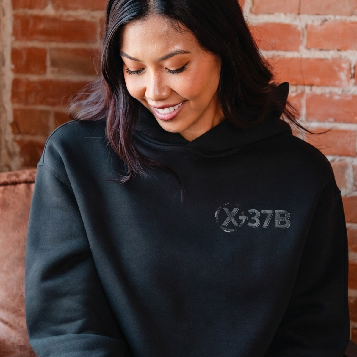A woman with long dark hair smiles and looks down while wearing a black BOEING X-37B Orbital Test Vehicle Unisex Graphic Hoodie, seated in front of a brick wall.