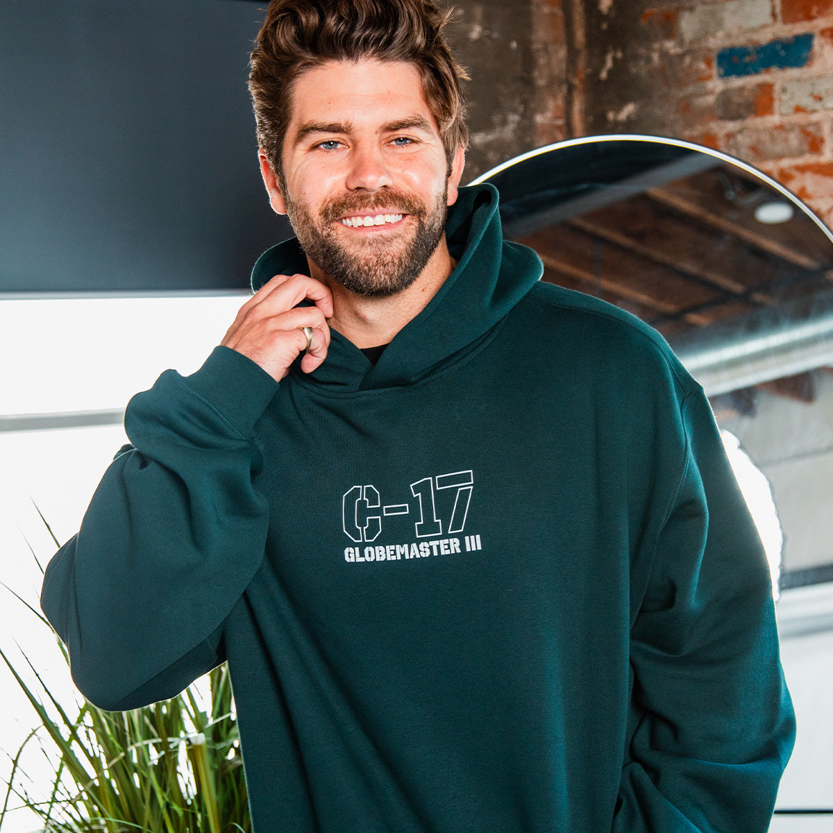 A bearded man smiles indoors near plants, wearing the BOEING C-17 Globemaster III Unisex Graphic Hoodie. Exposed brick and a round mirror are in the background—perfect for aviation enthusiasts.