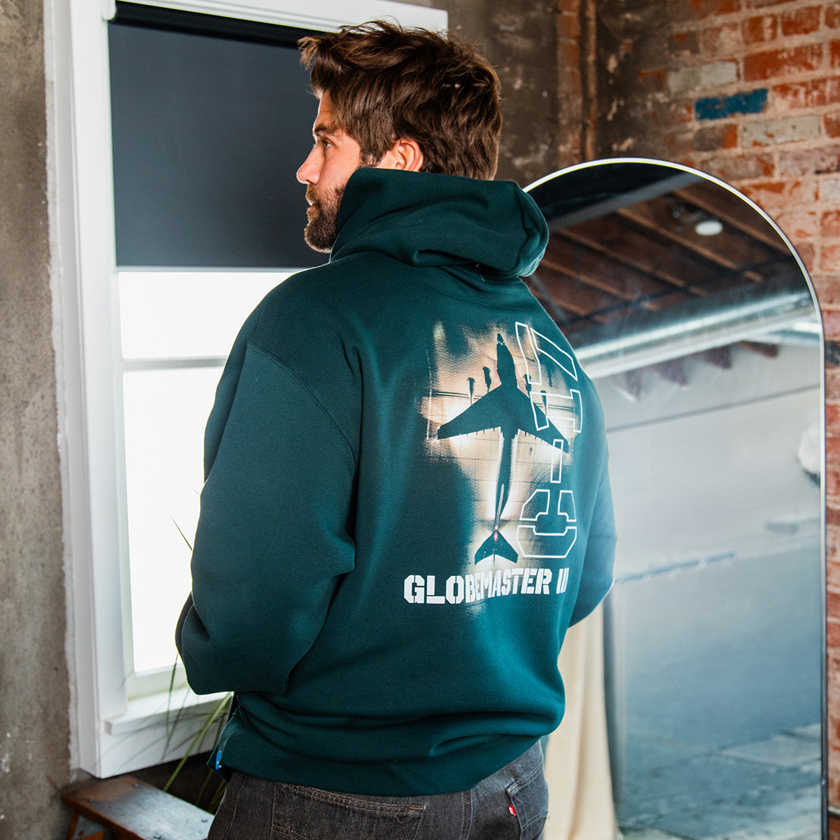 A man with a beard faces away indoors, wearing a green BOEING C-17 Globemaster III Unisex Graphic Hoodie with an airplane and "GLOBEMASTER III" on the back—ideal for aviation enthusiasts.