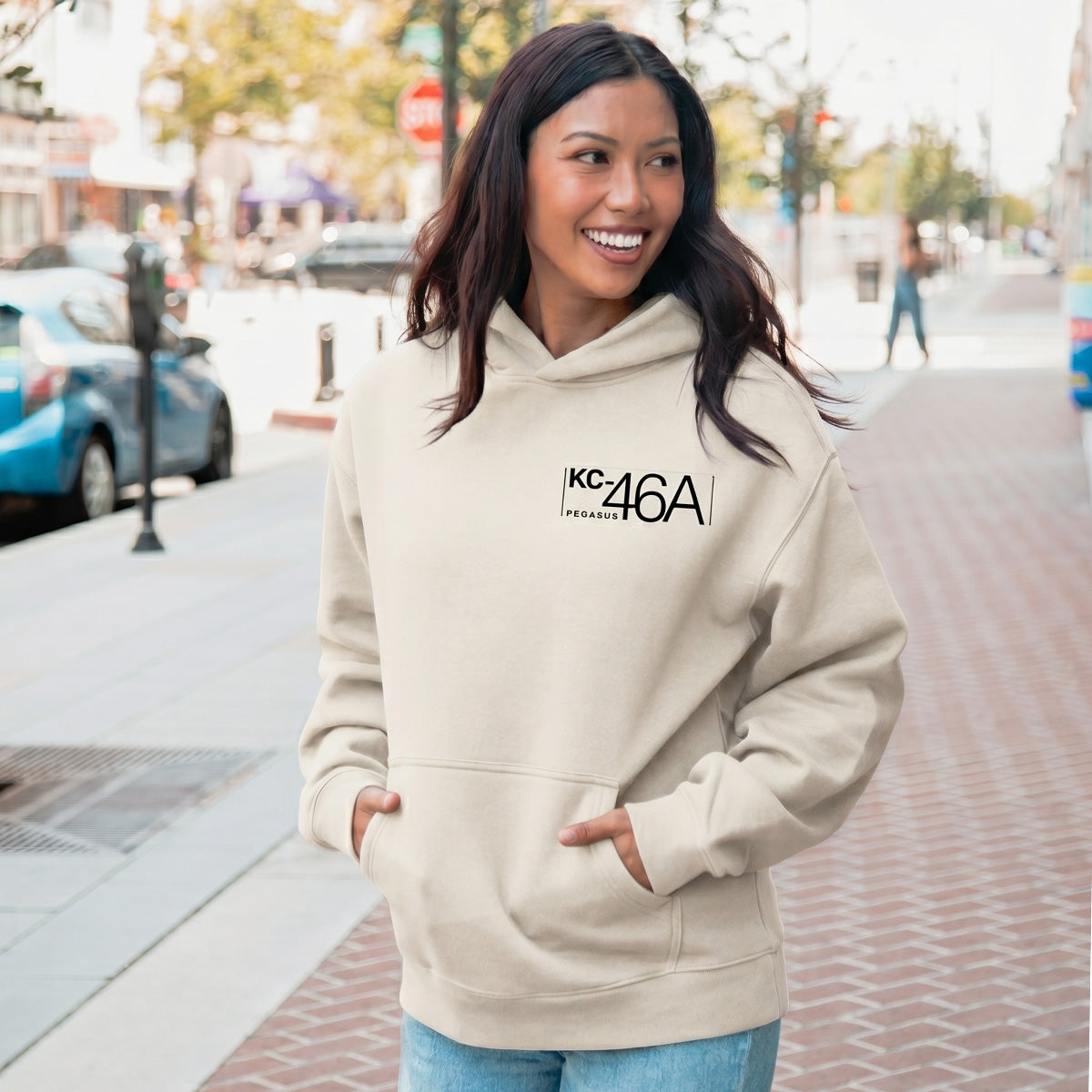 A woman smiles on a city sidewalk wearing the BOEING KC-46A Pegasus Unisex Graphic Hoodie—ideal for aviation fans. Parked cars, trees, and buildings fill the background.