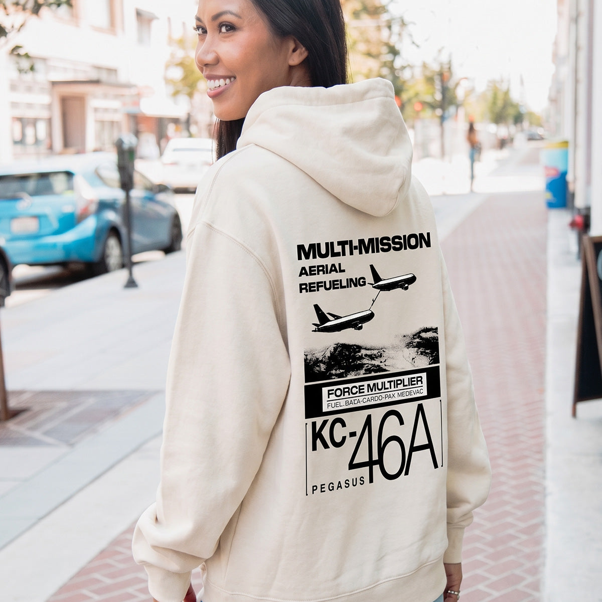 A smiling woman stands on a city sidewalk wearing a cream Boeing KC-46A Pegasus Unisex Graphic Hoodie by BOEING, featuring two airplanes and "MULTI-MISSION AERIAL REFUELING KC-46A PEGASUS" text—ideal for aviation fans.