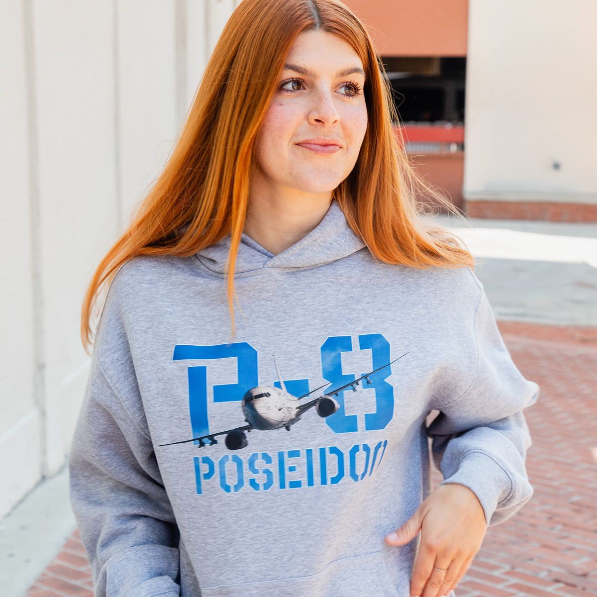 A woman with long red hair wears a BOEING P-8A Poseidon Unisex Graphic Hoodie while standing outdoors on a brick walkway next to a white building.