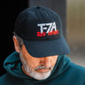 A man in a teal hoodie looks down while wearing the Boeing T-7A Red Hawk Graphic Hat by BOEING, featuring "T-7A RED HAWK" embroidered on the front—perfect for aviation enthusiasts.