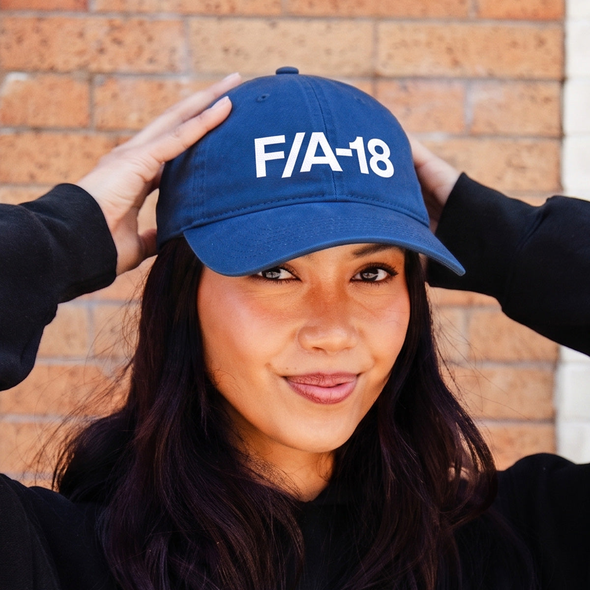 Aviation fans will love this image: a person in a black hoodie adjusts their BOEING F/A-18 Super Hornet Graphic Hat and smiles gently, with a brick wall behind them.