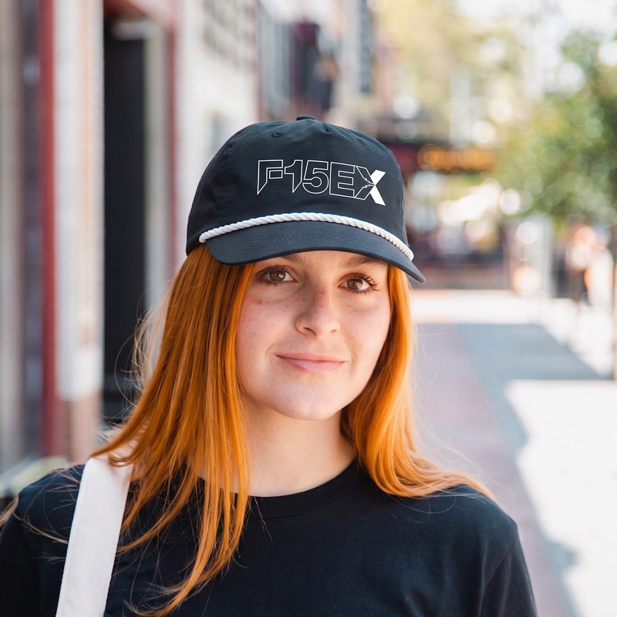 A young woman with long red hair wears a Boeing F-15EX Eagle II Graphic Hat by BOEING and a black t-shirt, standing outdoors on a sunny city street with blurred buildings and trees—ideal for aviation fans.