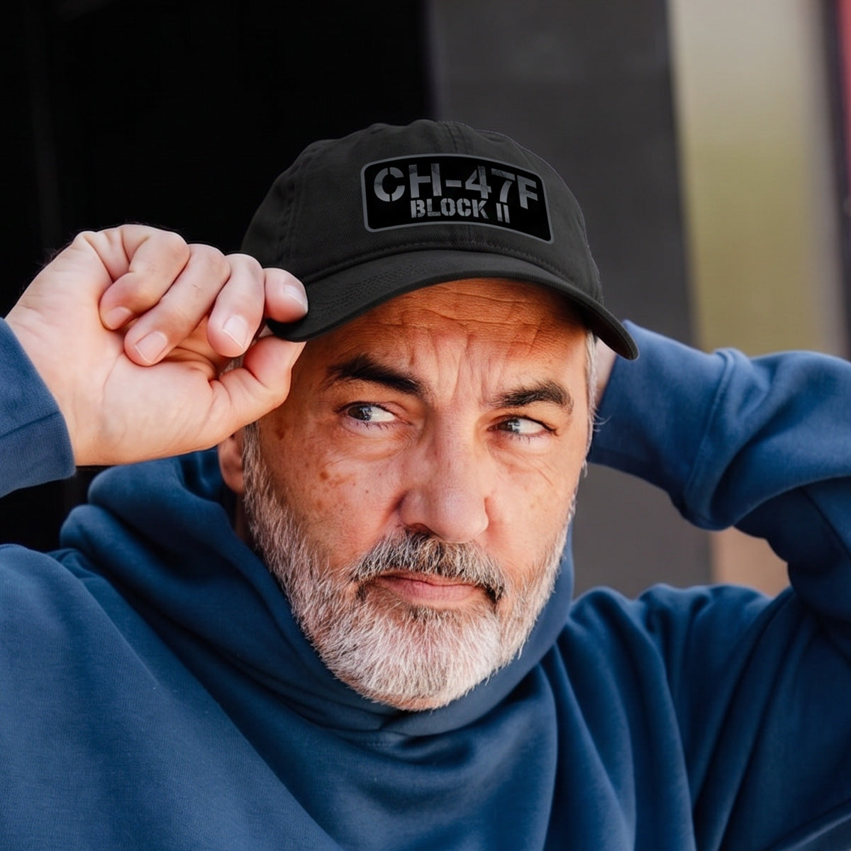 A man with a gray beard and mustache in a BOEING CH-47F Block II Graphic Hat and blue hoodie adjusts his cap, gazing thoughtfully to the side—an ideal look for aviation fans and Boeing supporters.