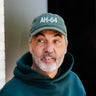 A man with a gray beard, in a dark green hoodie and BOEING AH-64 Apache Graphic Hat, looks right—ideal for aviation fans or Boeing supporters—standing against a dark background and white brick wall.