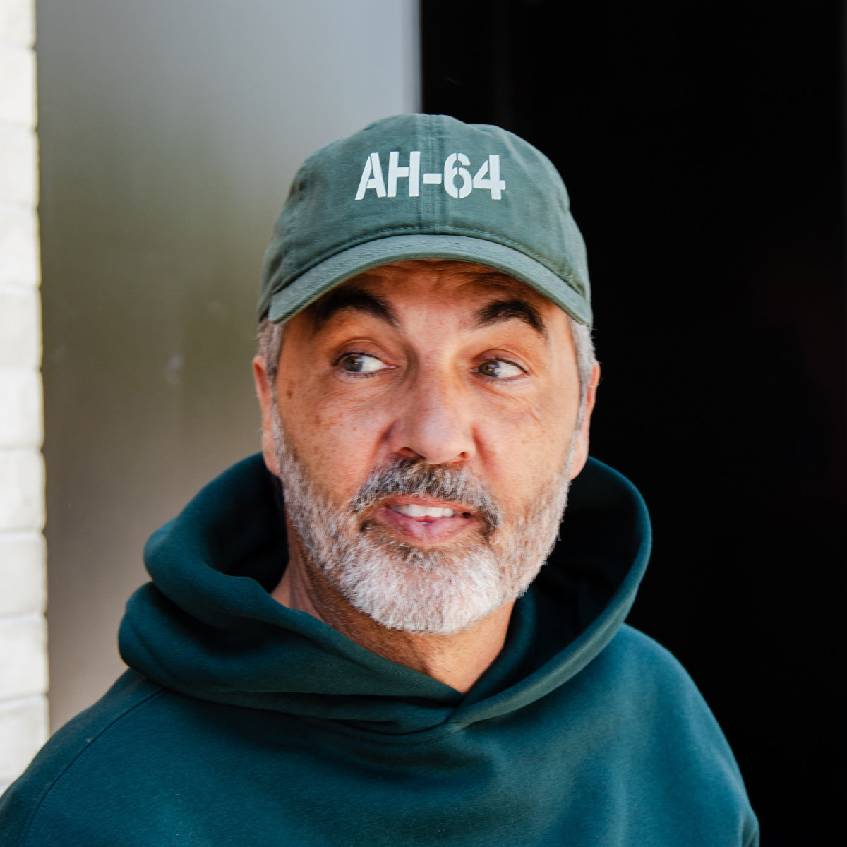 A man with a gray beard, in a dark green hoodie and BOEING AH-64 Apache Graphic Hat, looks right—ideal for aviation fans or Boeing supporters—standing against a dark background and white brick wall.