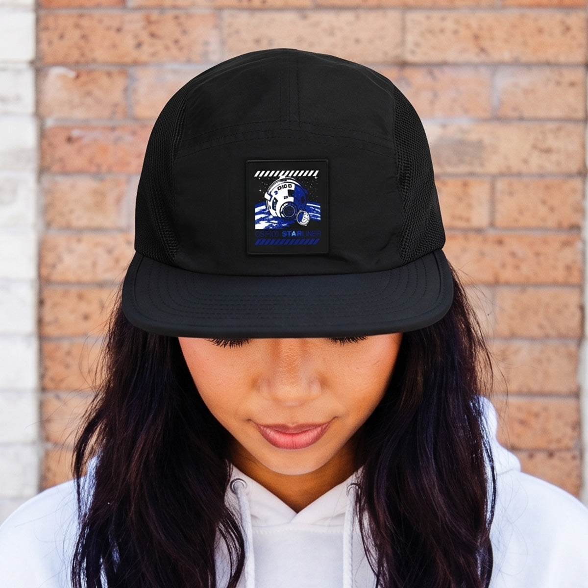 A woman stands against a brick wall, looking down, wearing the BOEING CST-100 Starliner Graphic Hat featuring a black design with a blue and white soccer ball patch.