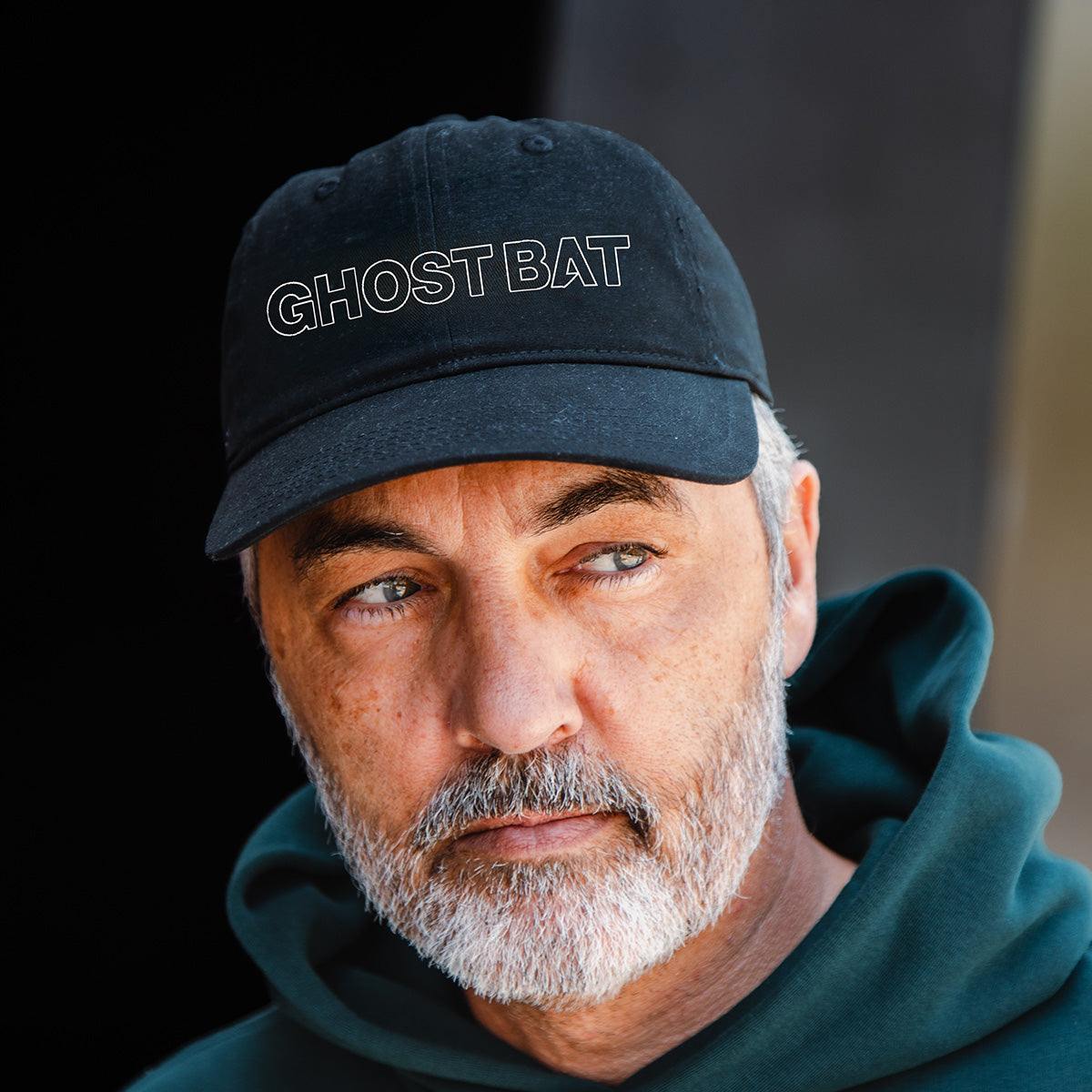 A middle-aged man with a gray beard wears a BOEING MQ-28 Ghost Bat Graphic Hat and a dark green hoodie, gazing thoughtfully to the side against a dark, out-of-focus background.