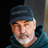 A middle-aged man with a gray beard wears a BOEING MQ-28 Ghost Bat Graphic Hat and a dark green hoodie, gazing thoughtfully to the side against a dark, out-of-focus background.