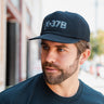 A bearded man in a black BOEING X-37B Orbital Test Vehicle Graphic Hat and matching tee stands outside on a sunny day, glancing left with a blurred backdrop of a building and trees.