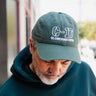 A man with a gray beard in a green hoodie and a BOEING C-17 Globemaster III Graphic Hat looks down while standing outdoors.