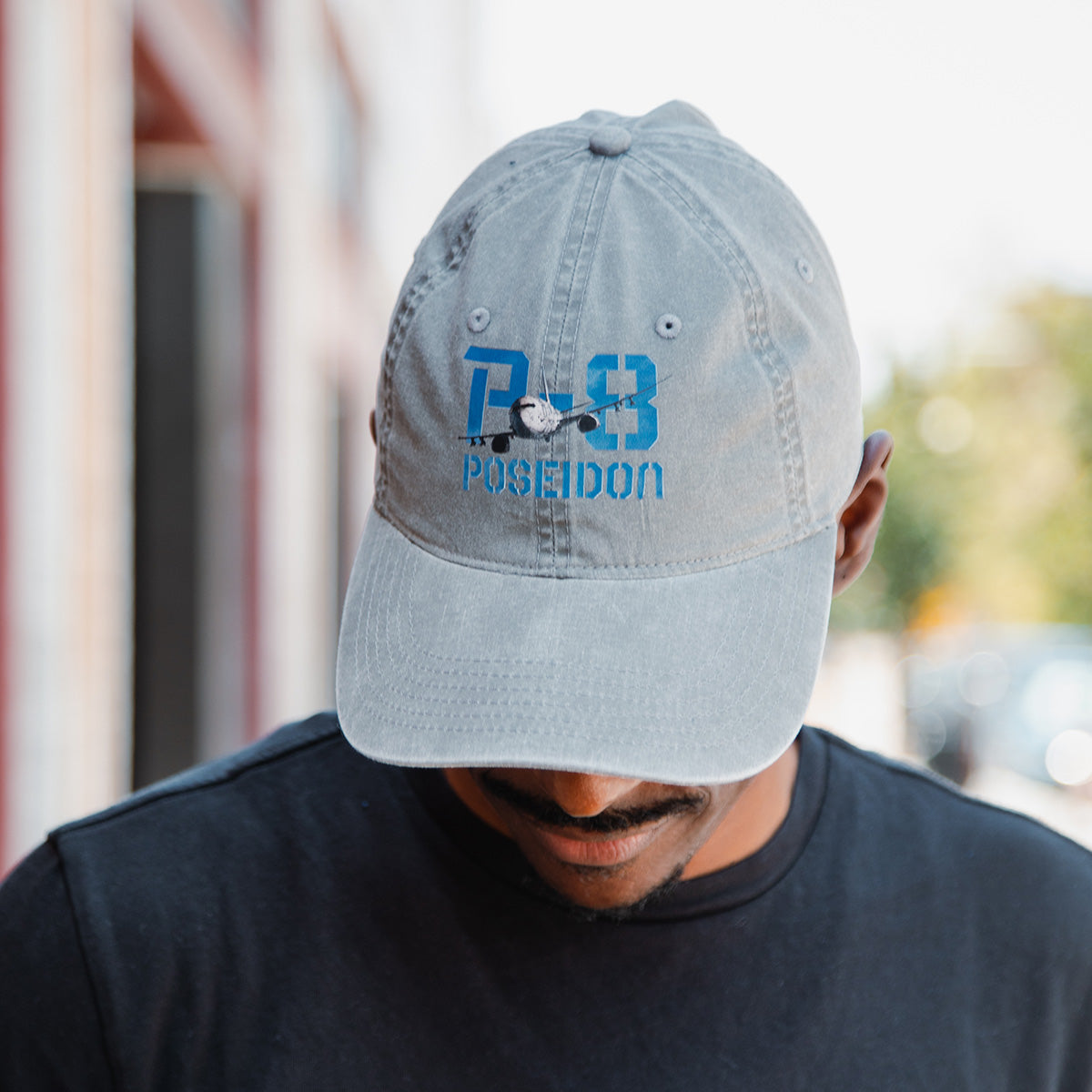 A person wears the BOEING P-8A Poseidon Graphic Hat in gray, featuring a blue aircraft design. This aviation enthusiast hat stands out as they look down, with buildings and greenery blurred in the background.