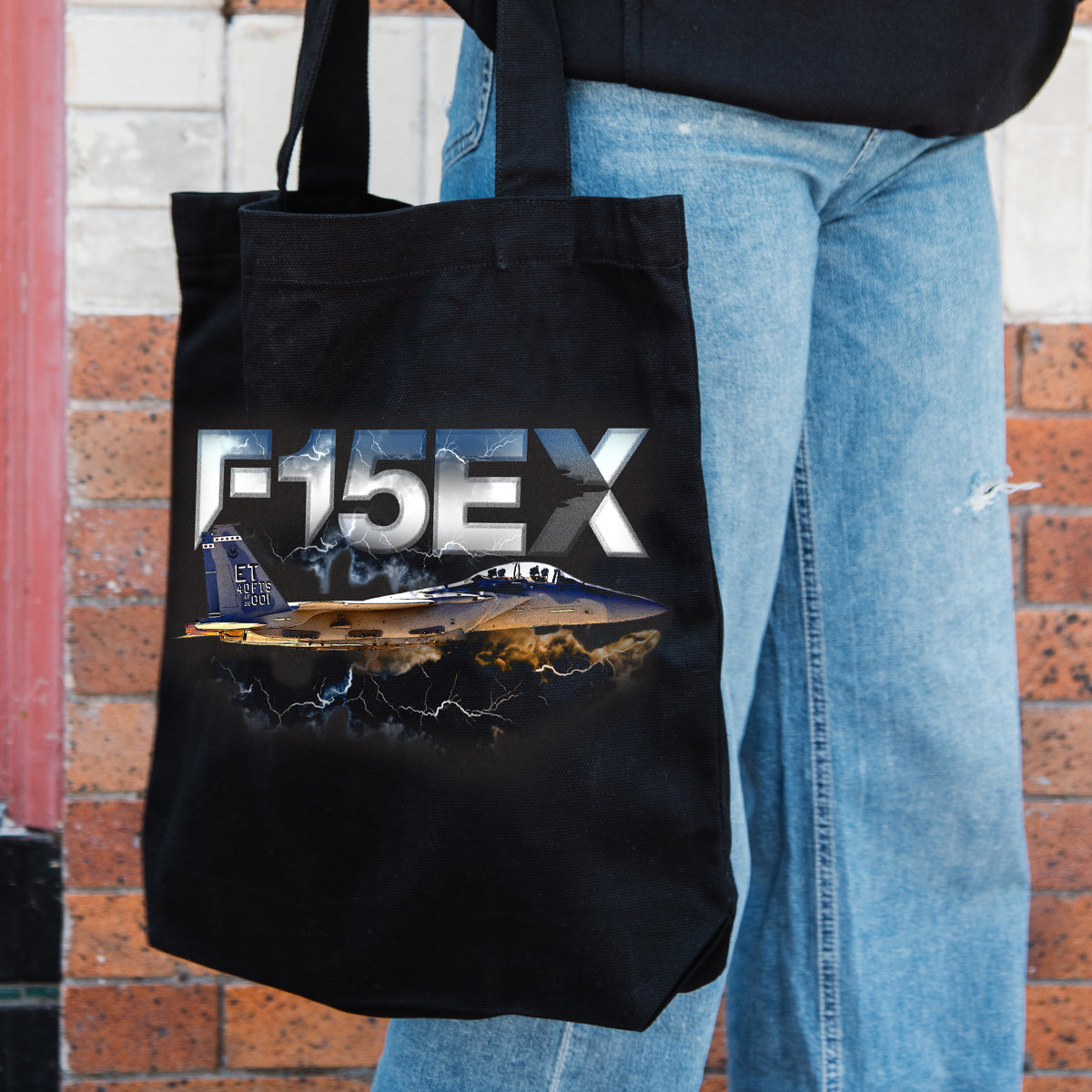 A person in blue jeans holds the BOEING F-15EX Eagle II Graphic Tote, showcasing a fighter jet with lightning and bold metallic F-15EX text—ideal for aviation enthusiasts.