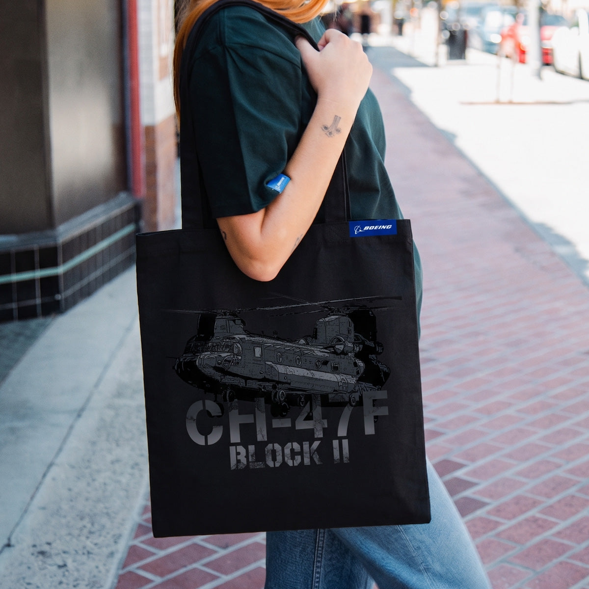 Carrying the Boeing CH-47F Block II Graphic Tote by BOEING, featuring a military helicopter design and blue Boeing tag, an aviation enthusiast walks along a brick sidewalk in the city.