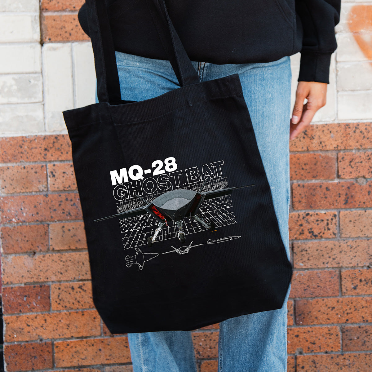 A person holds the BOEING MQ-28 Ghost Bat Graphic Tote, a black bag with a drone graphic—ideal for aviation fans. They're dressed in blue jeans and a black hoodie.