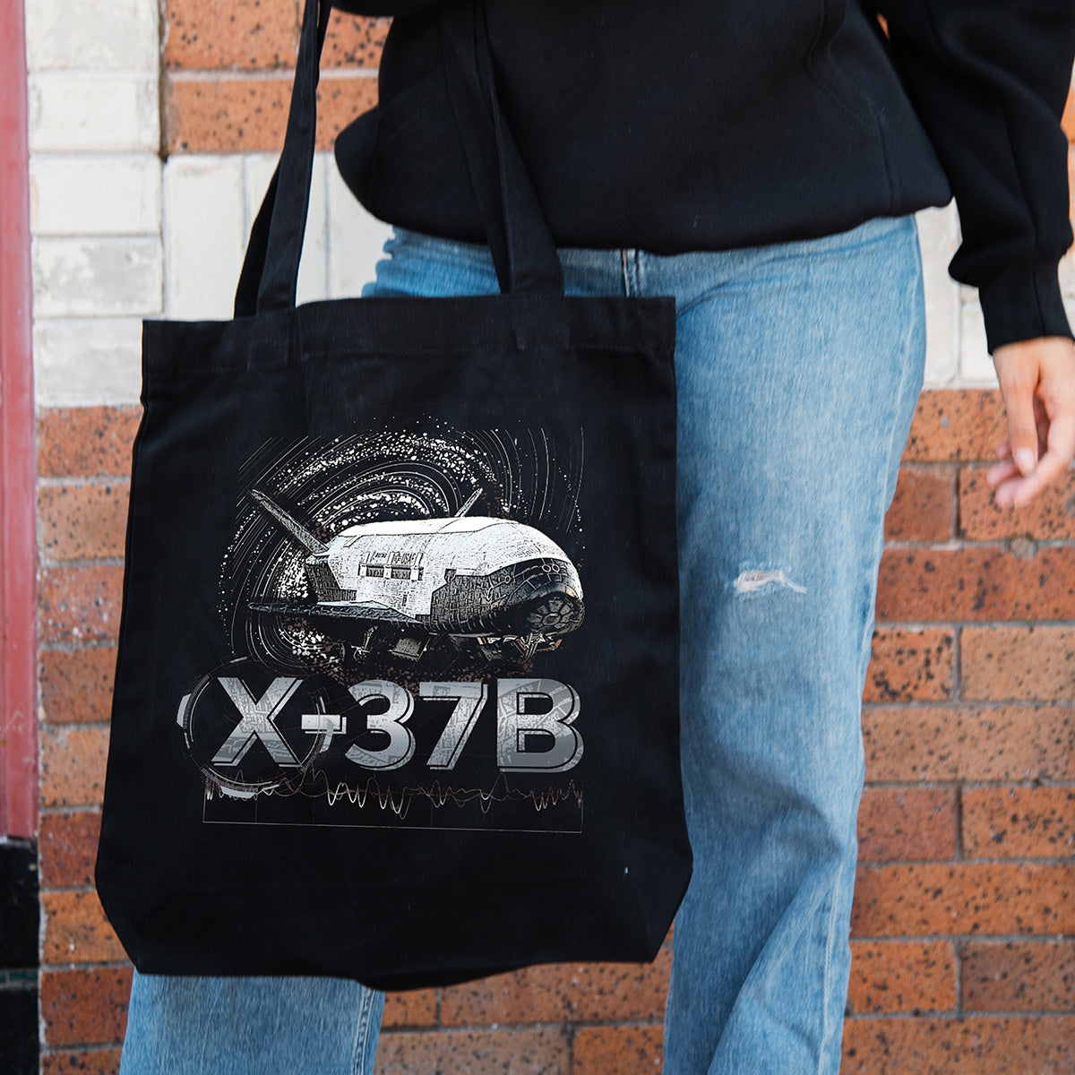 A person in blue jeans and a black sweater holds the BOEING X-37B Orbital Test Vehicle Graphic Tote, featuring detailed artwork of Boeing’s spaceplane. The scene is set against a brick wall background.