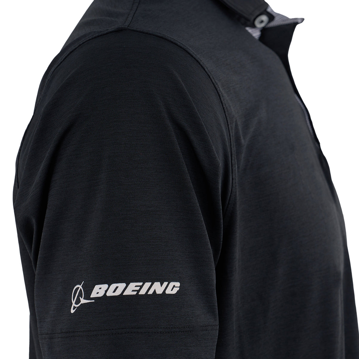 A close-up shows a person wearing the Boeing Space Launch System Series Men’s Polo by BOEING. The black polo features a white Boeing logo on the left sleeve and moisture-wicking fabric for comfort. Only the upper torso and part of the arm are visible.