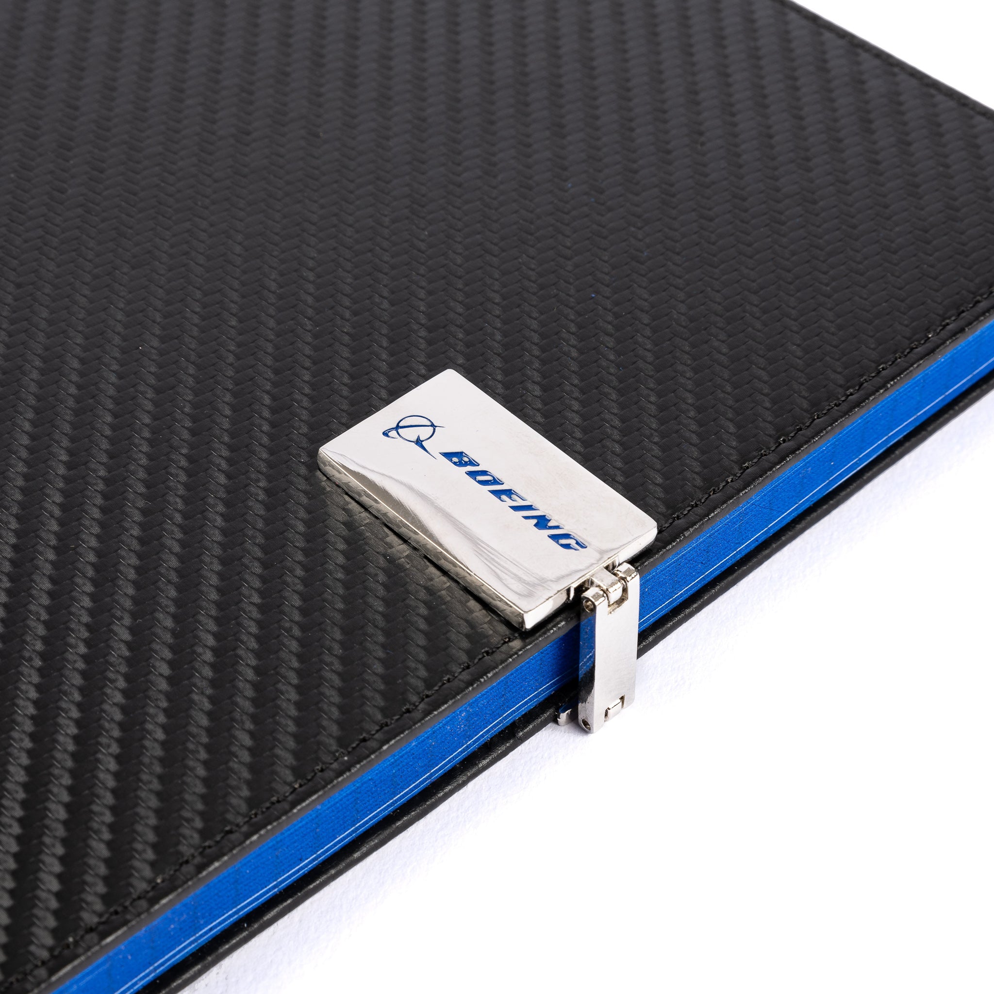 A close-up of the Boeing Carbon Fiber Notebook from The Boeing Store features a sleek black textured design with a carbon fiber cover and metallic hinge. The Boeing logo is on a silver metal plate, with blue-edged highlights against a white background.