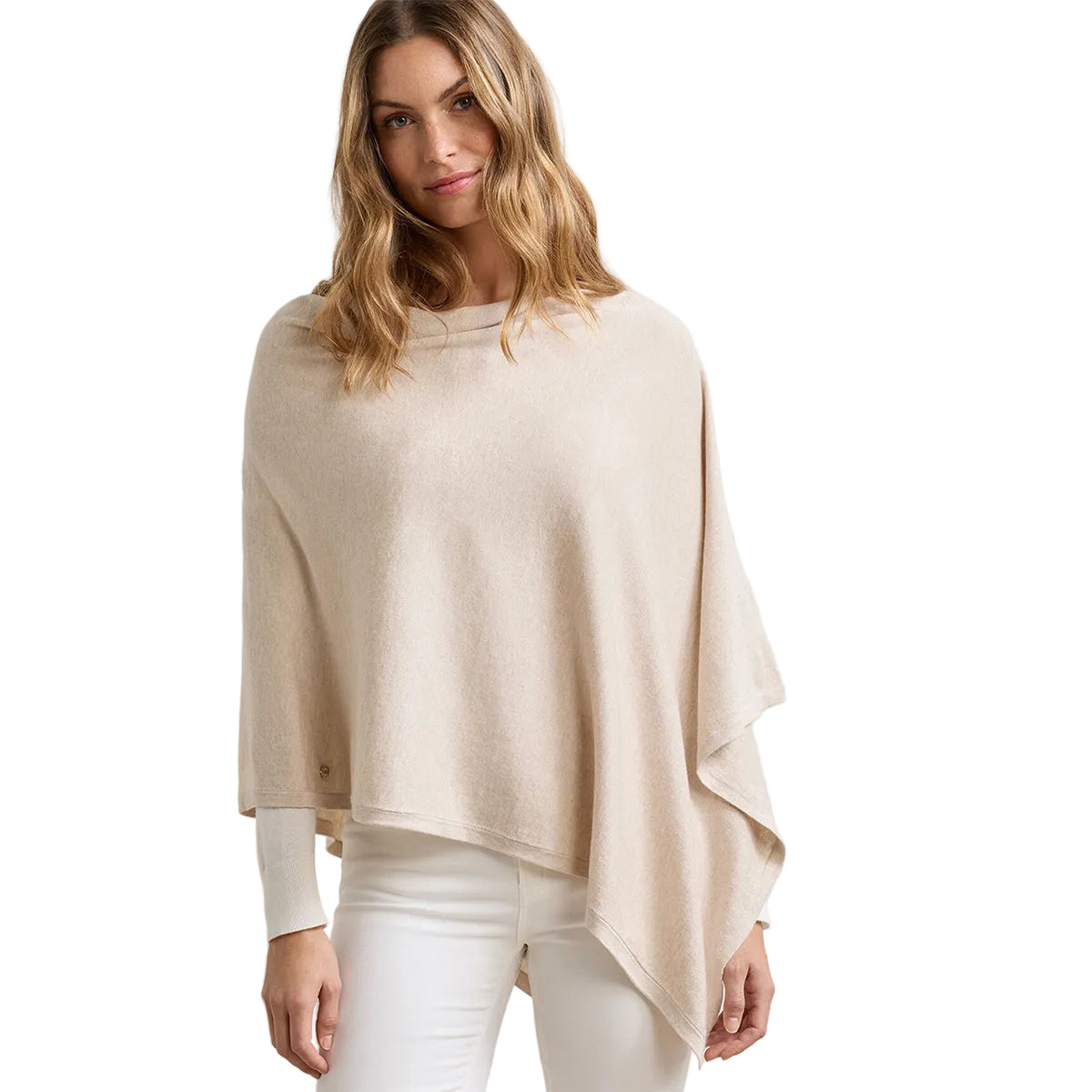 A woman with wavy blonde hair models the beige Peter Millar Boeing Women’s Spring Blend Poncho by BOEING over a white long-sleeve top and white pants, standing against a plain white background.