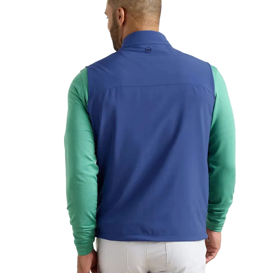 A man is seen from the back wearing a blue Peter Millar Boeing Men’s Dunes Performance Vest over a green long-sleeve shirt and light-colored pants, standing against a plain white background.