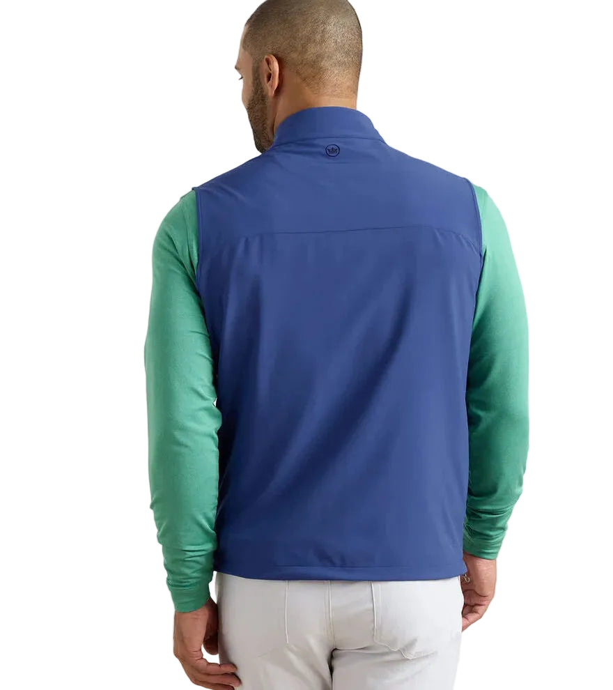 A man is seen from the back wearing a blue Peter Millar Boeing Men’s Dunes Performance Vest over a green long-sleeve shirt and light-colored pants, standing against a plain white background.